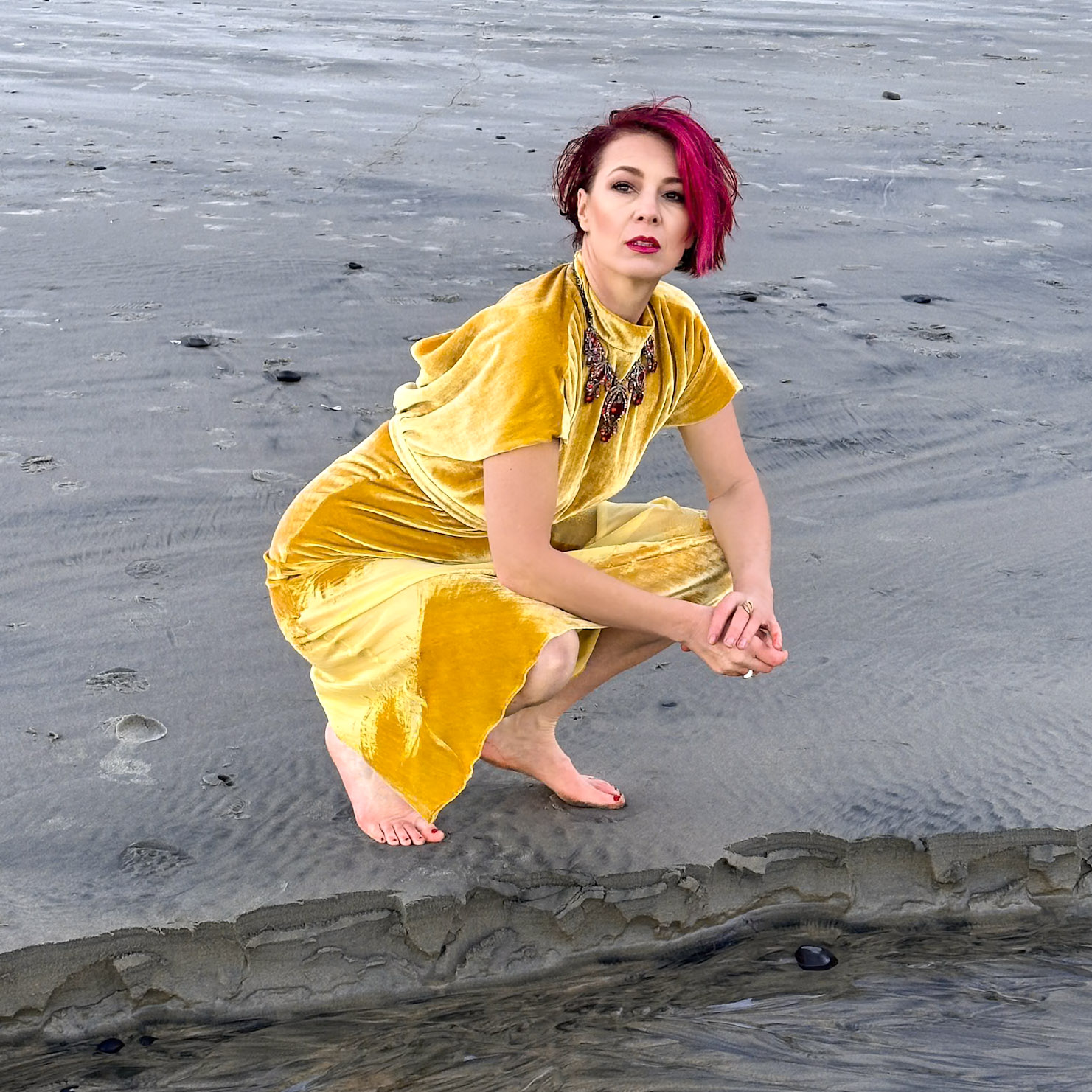 Viktoria Sway in a gold velvet dress, crouched barefoot on grey sand, steady direct gaze